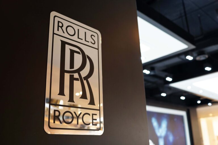 Primer plano del logotipo de Rolls-Royce sobre una pared oscura, con un entorno interior iluminado al fondo.