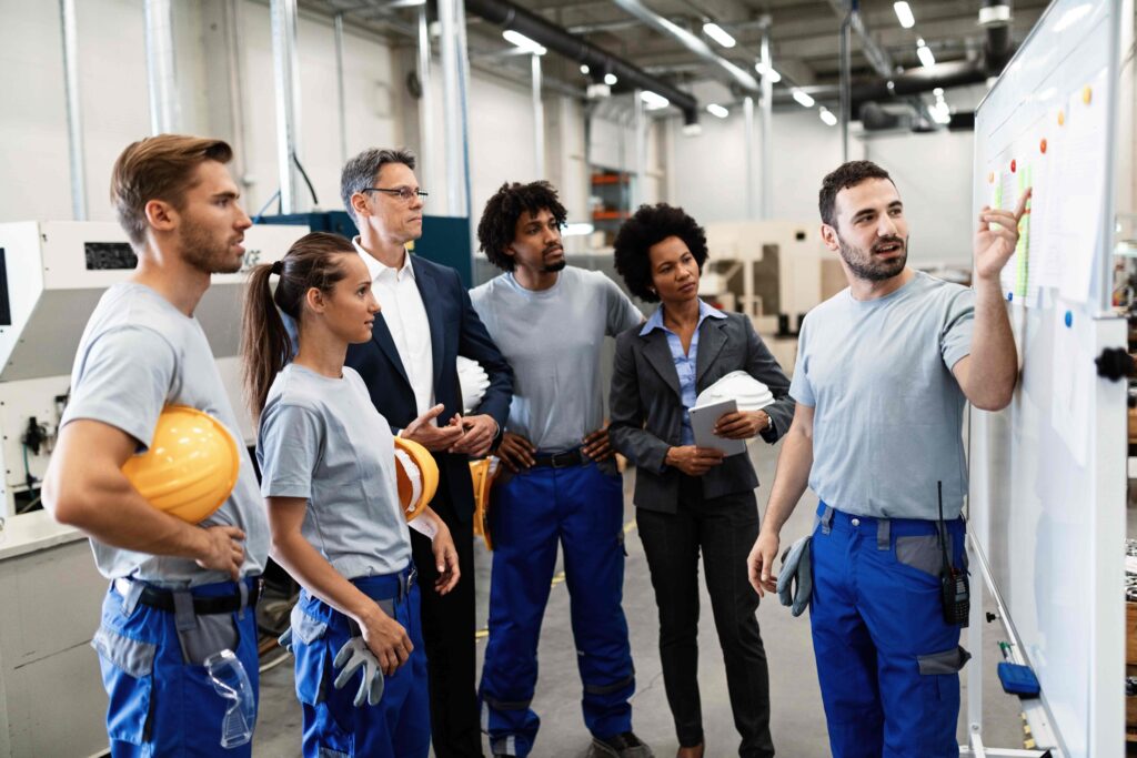 Equipo de operarios y gerentes reunidos frente a un tablero en un taller de fabricación, analizando métricas y estrategias de producción.