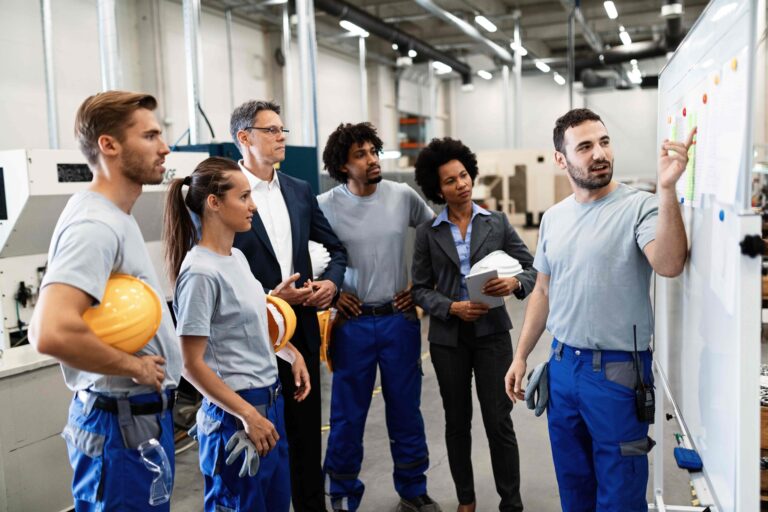 Equipo de operarios y gerentes reunidos frente a un tablero en un taller de fabricación, analizando métricas y estrategias de producción.