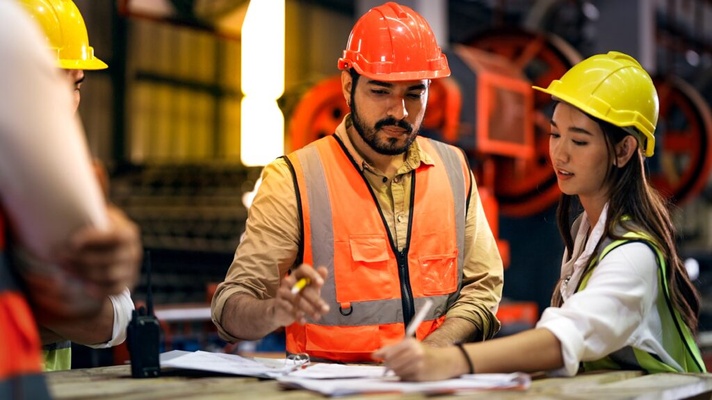 Supervisor y trabajadora con cascos de seguridad revisando planos y documentos en un taller de fabricación.