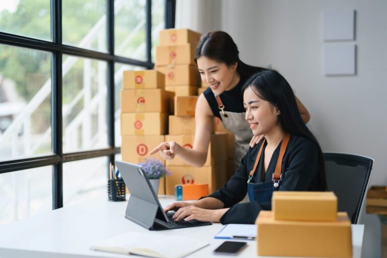 Dos mujeres emprendedoras gestionan ventas en línea rodeadas de cajas, ilustrando cómo las empresas jóvenes generan empleo y promueven productividad e innovación.
