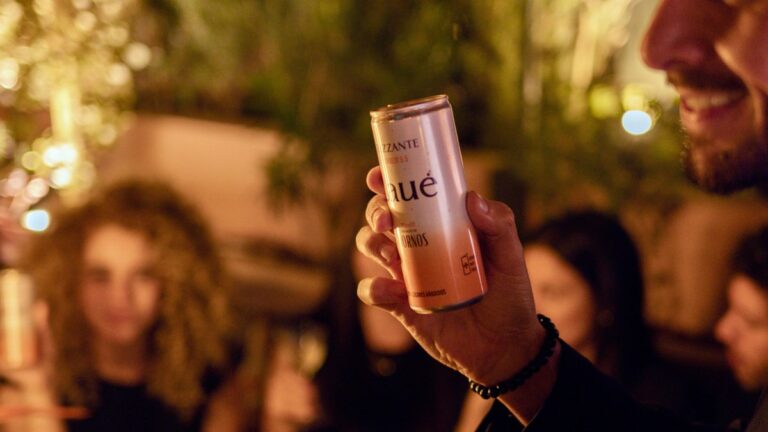 Mano de un invitado sosteniendo una lata de Laué Frizzante en un ambiente nocturno con luces cálidas y personas celebrando en segundo plano.