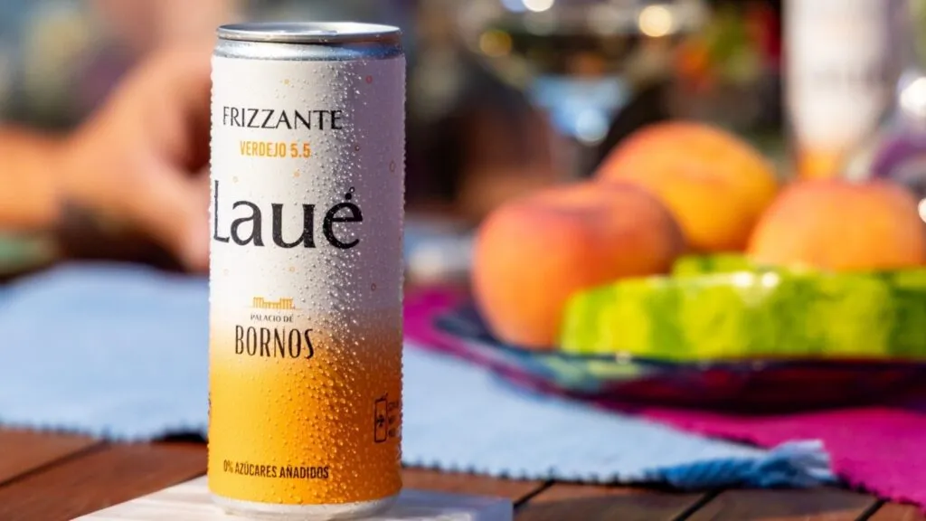Lata de LAUÉ frizzante bien fría con gotas de condensación, colocada sobre una base blanca en una mesa de madera. Al fondo se observan frutas frescas, un mantel colorido y copas desenfocadas que sugieren un momento de convivencia al aire libre.