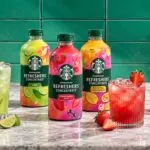 Botellas de concentrados frutales acompañadas por vasos con bebidas coloridas, hielo y frutas frescas en una cocina moderna.