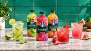 Botellas de concentrados frutales acompañadas por vasos con bebidas coloridas, hielo y frutas frescas en una cocina moderna.