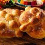 Pan de muerto tradicional mexicano cubierto de azúcar y decorado con figuras de huesos, acompañado de flores de cempasúchil.