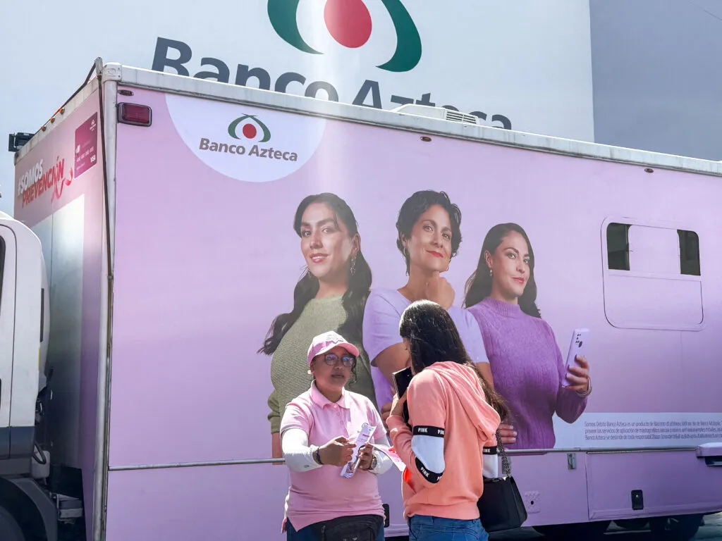 Promotora de Banco Azteca conversa con una mujer frente a una unidad móvil rosa de la campaña SOMOS Prevención.
